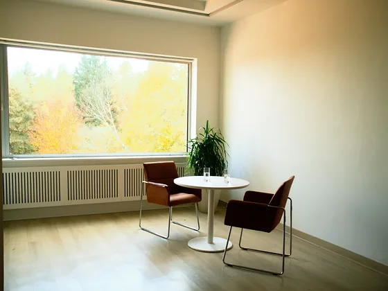 A small round table with two glasses of water between two chairs, beside a window onto autumn trees