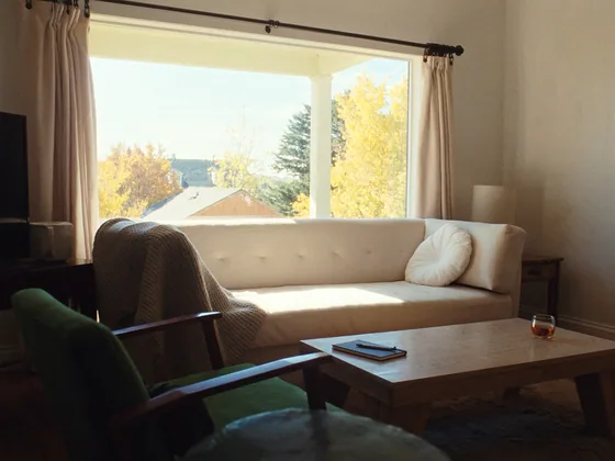 A quiet living room corner with a sofa, a throw and a closed notebook on the coffee table, autumn trees outside